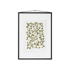 A framed botanical art featuring pressed green leaves on a white sheet of paper, enclosed in a black frame with a hanging cord, shown against a plain white background.