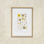 A framed botanical art print featuring pressed flowers and leaves on textured white paper, set in a oak wood frame against a light beige wall.