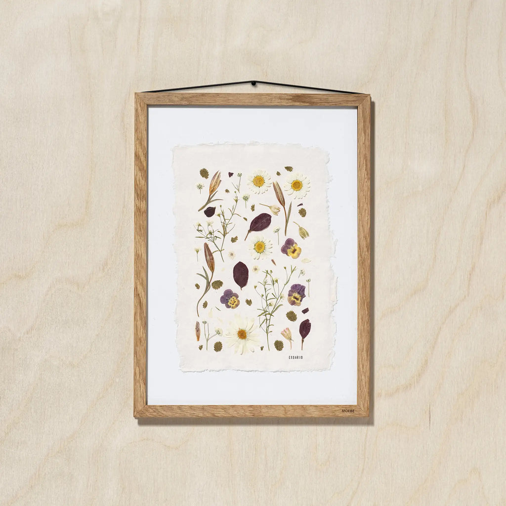 A framed botanical art featuring delicate pressed flowers on a textured off-white sheet of paper, mounted with a white background and a light oak wood frame, hanging on a light oak wood wall.