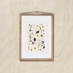 A framed botanical art featuring delicate pressed flowers on a textured off-white sheet of paper, mounted with a white background and a light oak wood frame, hanging on a light oak wood wall.