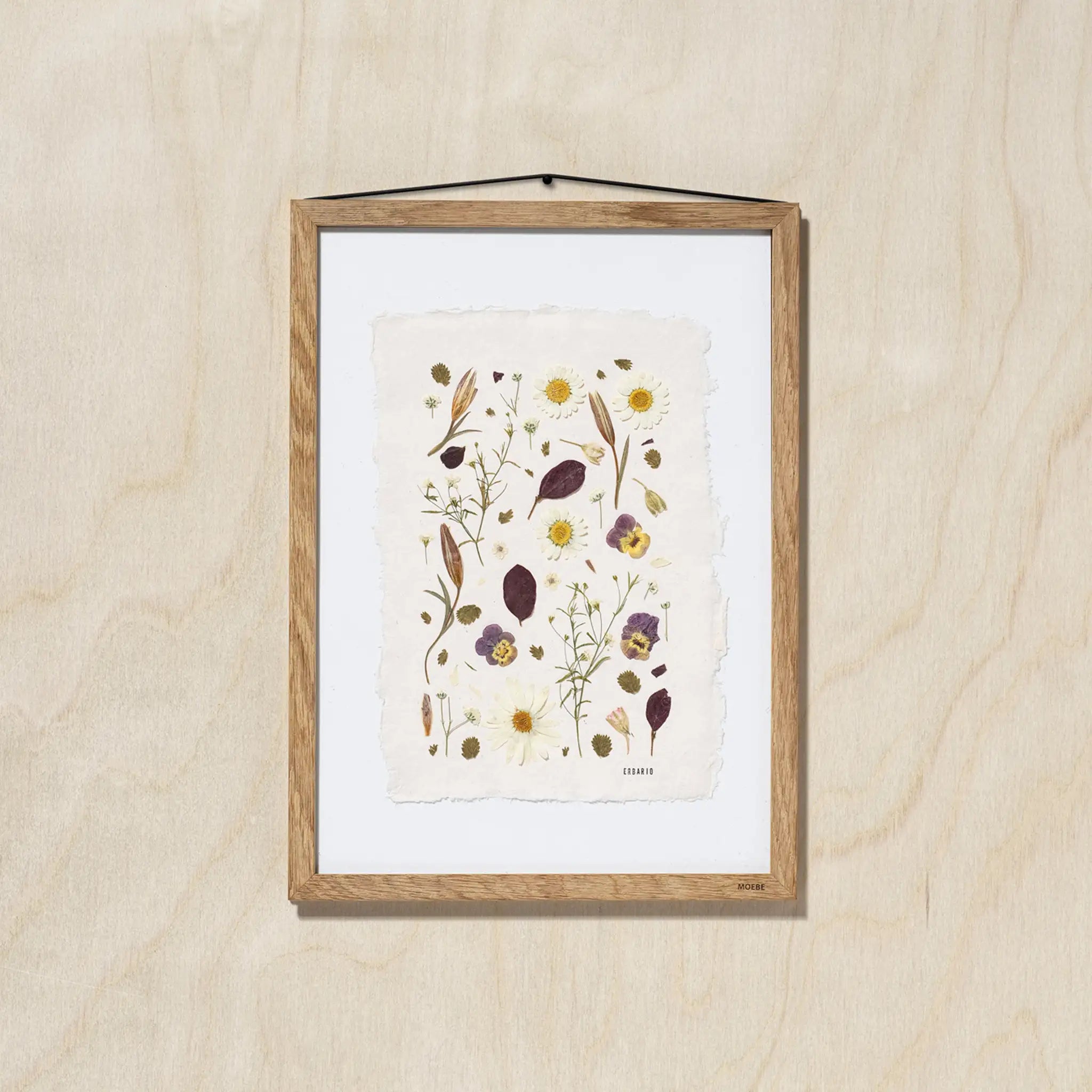A framed botanical art featuring delicate pressed flowers on a textured off-white sheet of paper, mounted with a white background and a light oak wood frame, hanging on a light oak wood wall.
