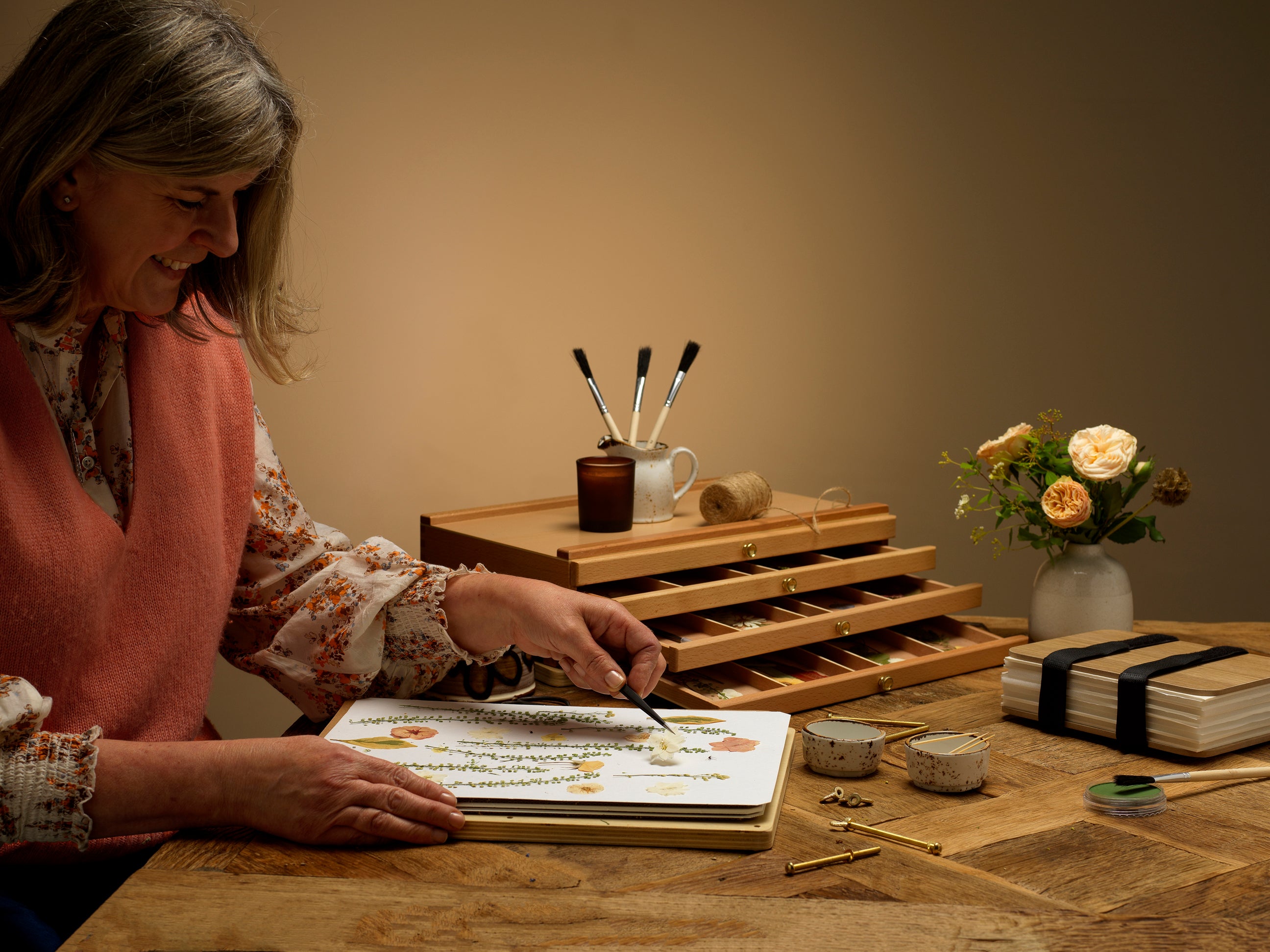 A woman sat at a wooden table, making pressed flower art work. The scene includes a wooden multi-compartment organiser, small ceramic containers, a stack of books tied with a black band, and a vase of flowers against a warm beige background.