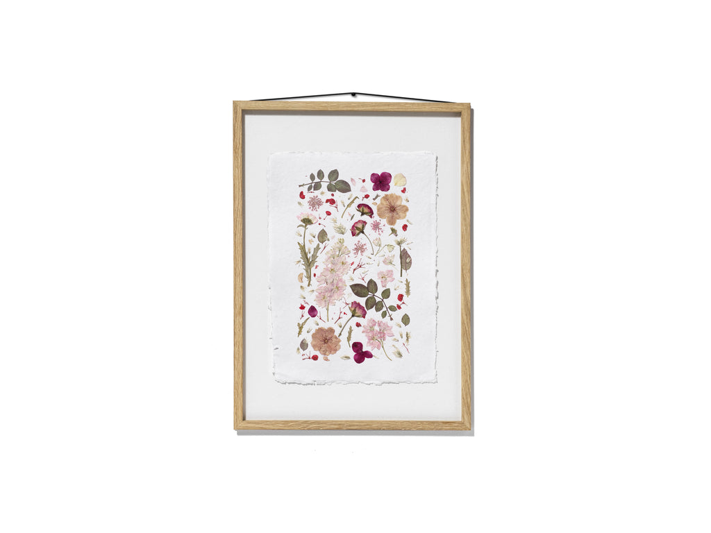 A framed botanical art featuring delicate pressed flowers and leaves in soft pink, burgundy, beige, and olive green tones. The print is mounted on a white textured sheet and enclosed in a light natural wood frame with a black hanging cord, shown against a solid white background.