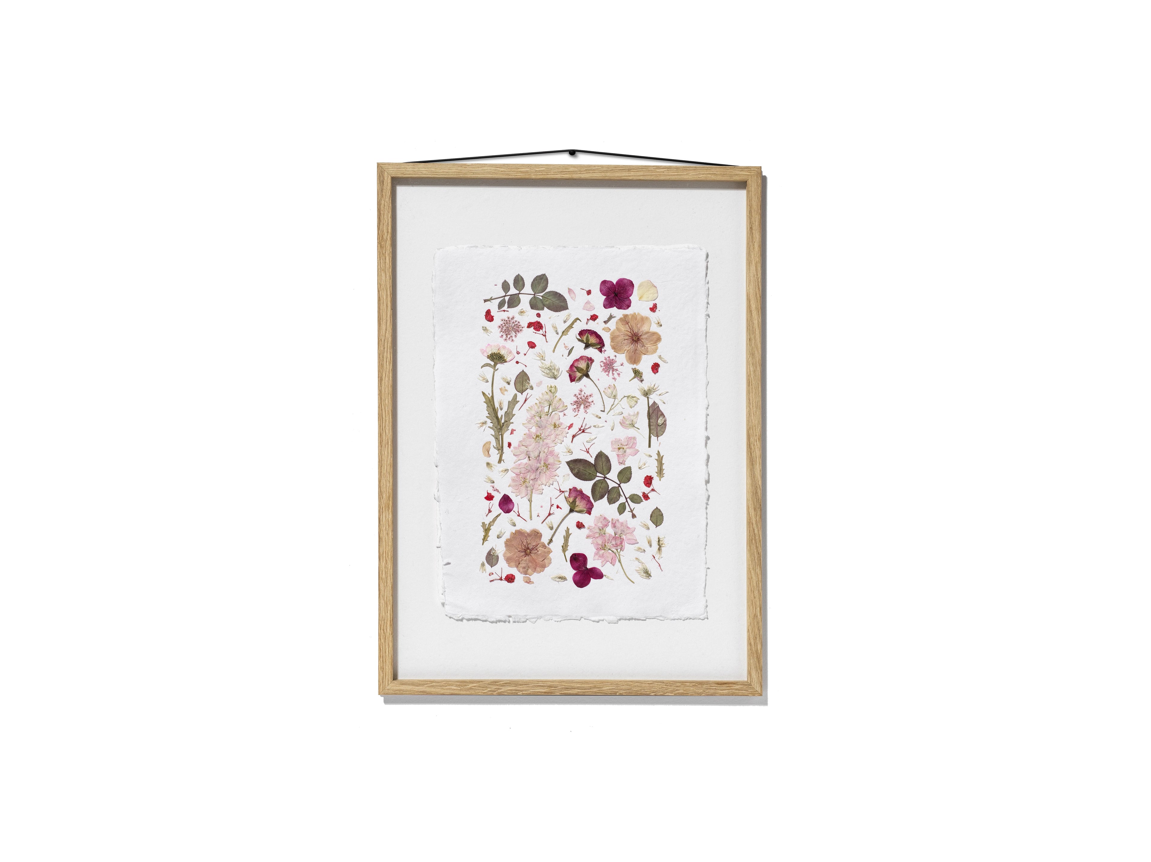 A framed botanical art featuring delicate pressed flowers and leaves in soft pink, burgundy, beige, and olive green tones. The print is mounted on a white textured sheet and enclosed in a light natural wood frame with a black hanging cord, shown against a solid white background.