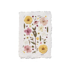 A square white paper art with irregular deckled edges, featuring a botanical illustration of pressed flowers in yellow, pink, and white with green leaves, shown on a plain white background.