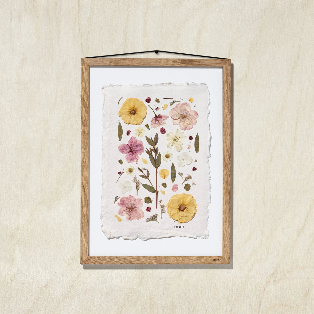 A framed botanical art featuring pressed flowers and leaves on a textured off-white sheet, mounted in a oak wood frame against a light oak wall.