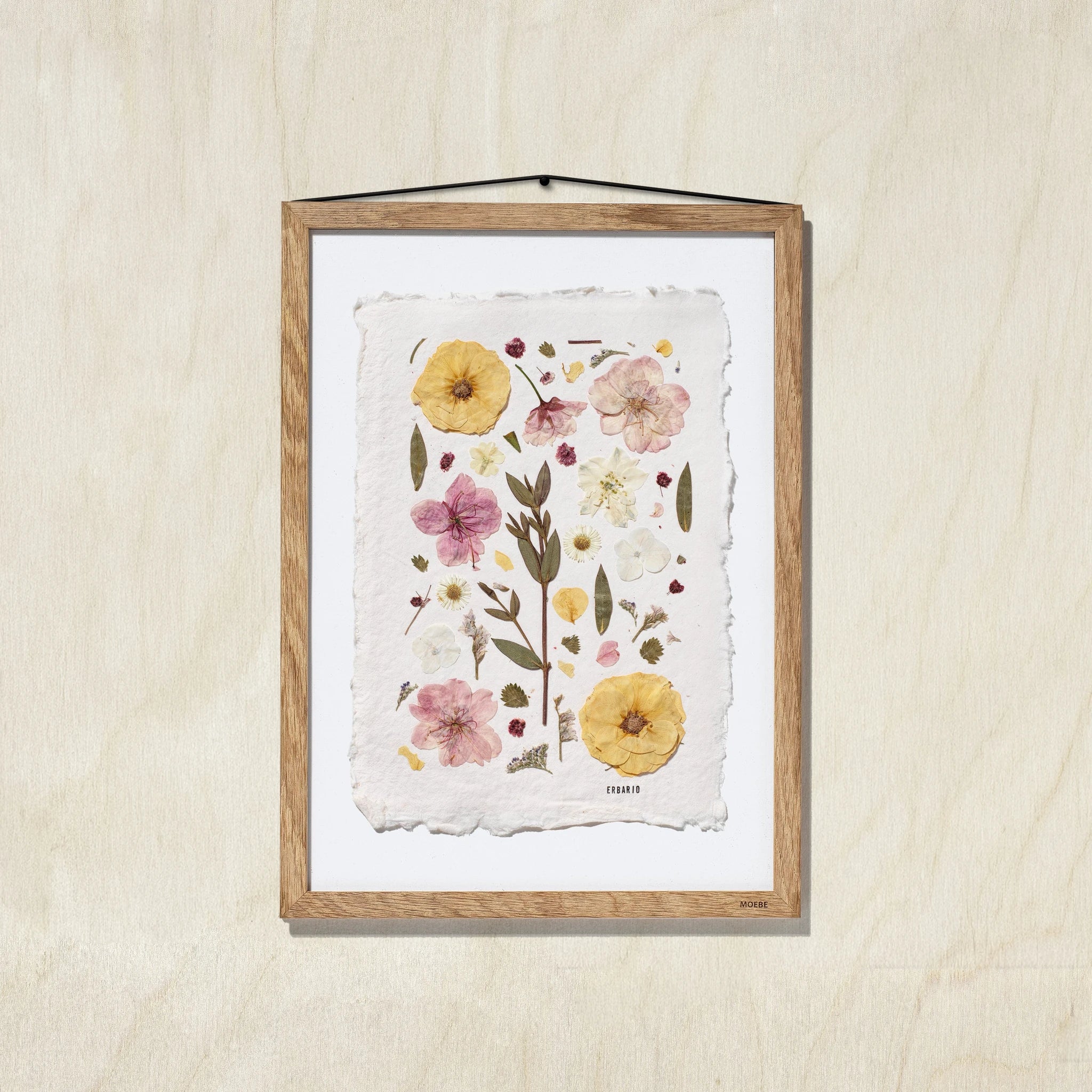 A framed botanical art featuring pressed flowers and leaves on a textured off-white sheet, mounted in a oak wood frame against a light oak wall.