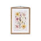 Framed botanical art featuring pressed flowers and leaves on textured off-white paper, housed in a natural oak wood frame with a black hanging string, shown against a plain white background.