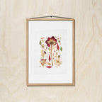 A framed botanical art featuring pressed flower and leaves, hung on a light wood wall with a black hanging string.