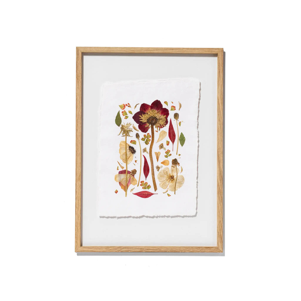 Framed botanical art featuring pressed flowers and leaves on textured white paper, set in a light natural wood frame against a white background.