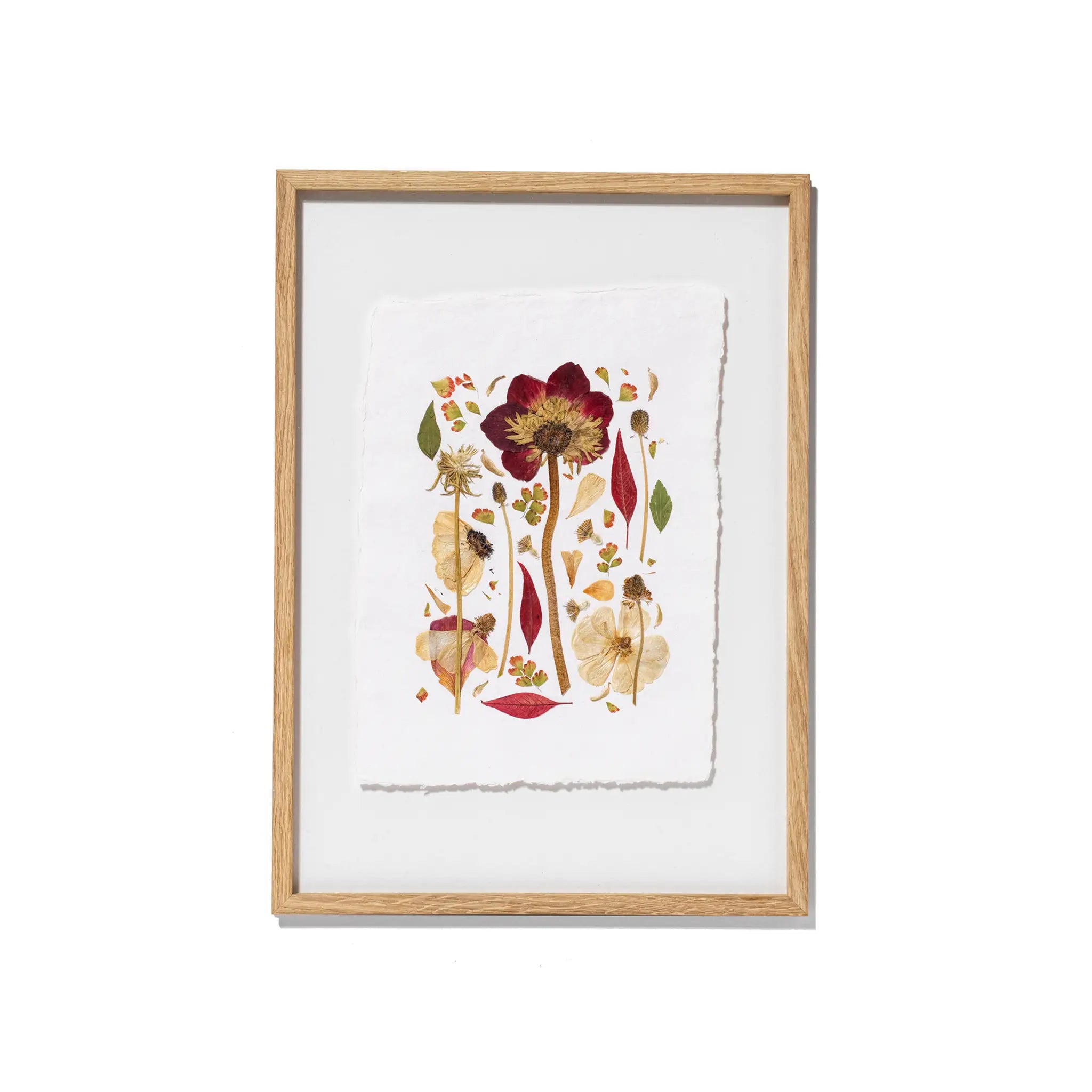 Framed botanical art featuring pressed flowers and leaves on textured white paper, set in a light natural wood frame against a white background.