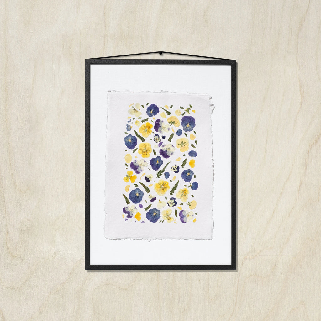 A framed floral art print featuring purple, blue and yellow pansies on a textured white sheet of paper, in a black frame hung against a light wood background.