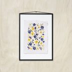 A framed floral art print featuring purple, blue and yellow pansies on a textured white sheet of paper, in a black frame hung against a light wood background.