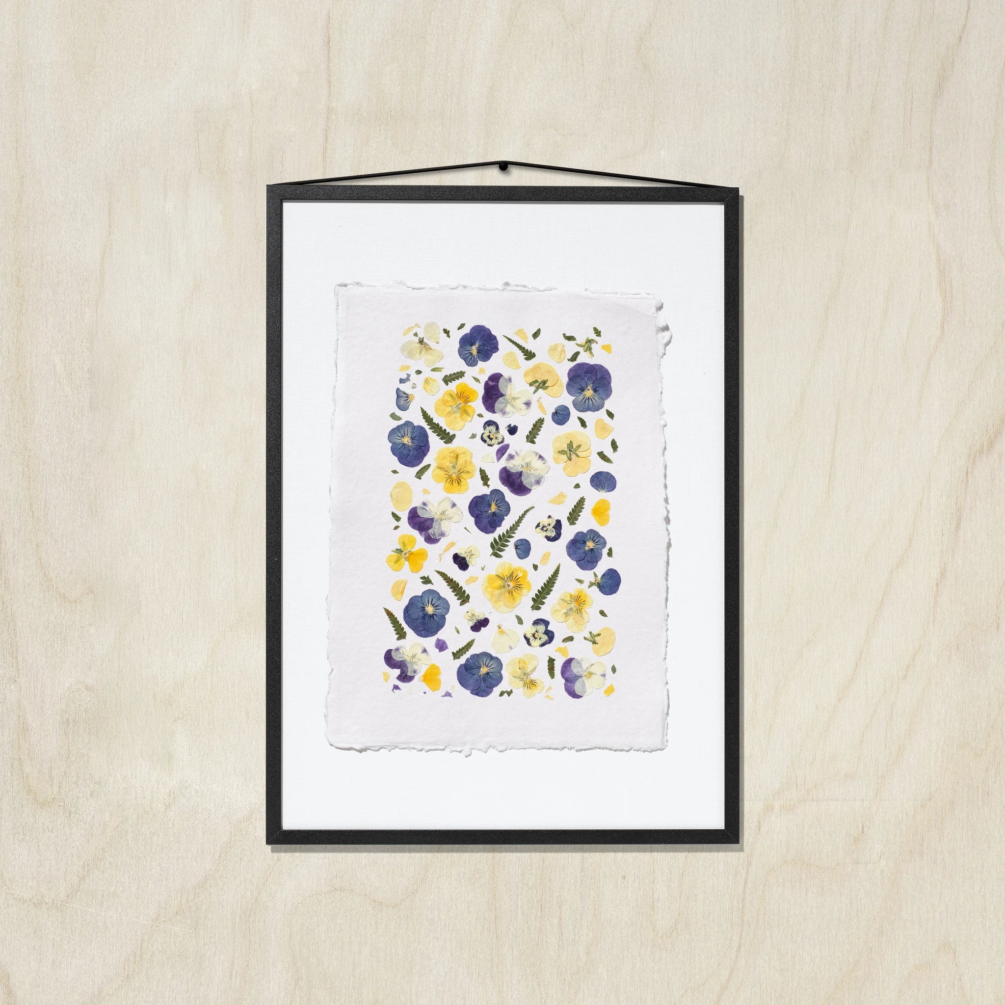 A framed floral art print featuring purple, blue and yellow pansies on a textured white sheet of paper, in a black frame hung against a light wood background.