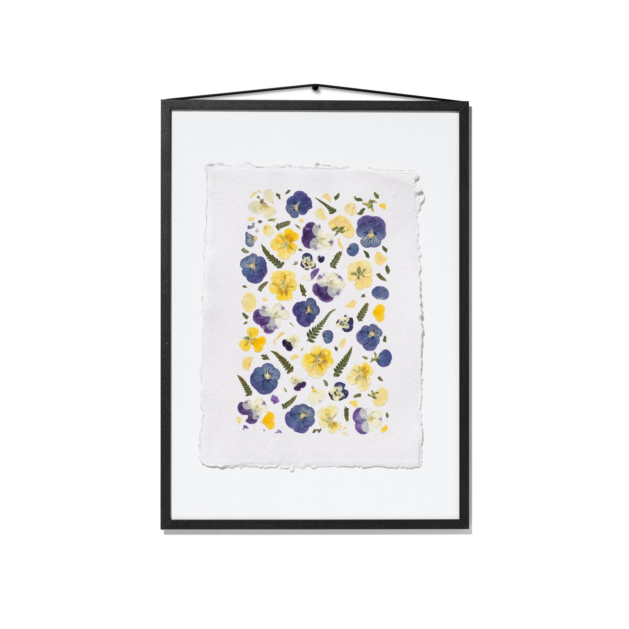 A framed floral art print featuring purple, blue and yellow pressed pansies on a textured white sheet of paper in a black frame, hung on a white background.