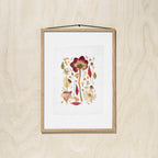 A framed botanical print featuring a pressed flower and leaves in muted red, beige, and green tones, mounted on a light wood wall with a black hanging string.