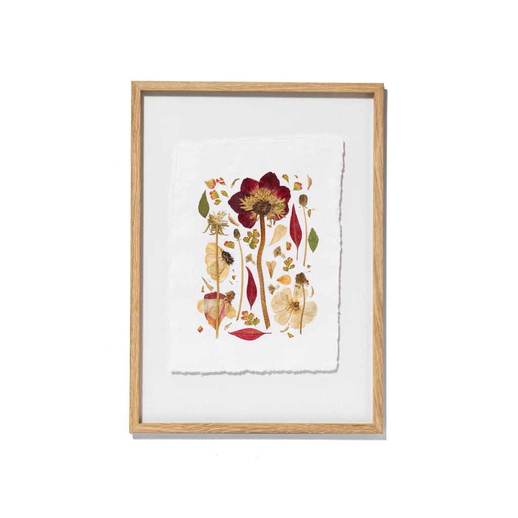 Framed botanical art featuring pressed flowers and leaves on textured white paper, set in a light natural wood frame against a clean white background.