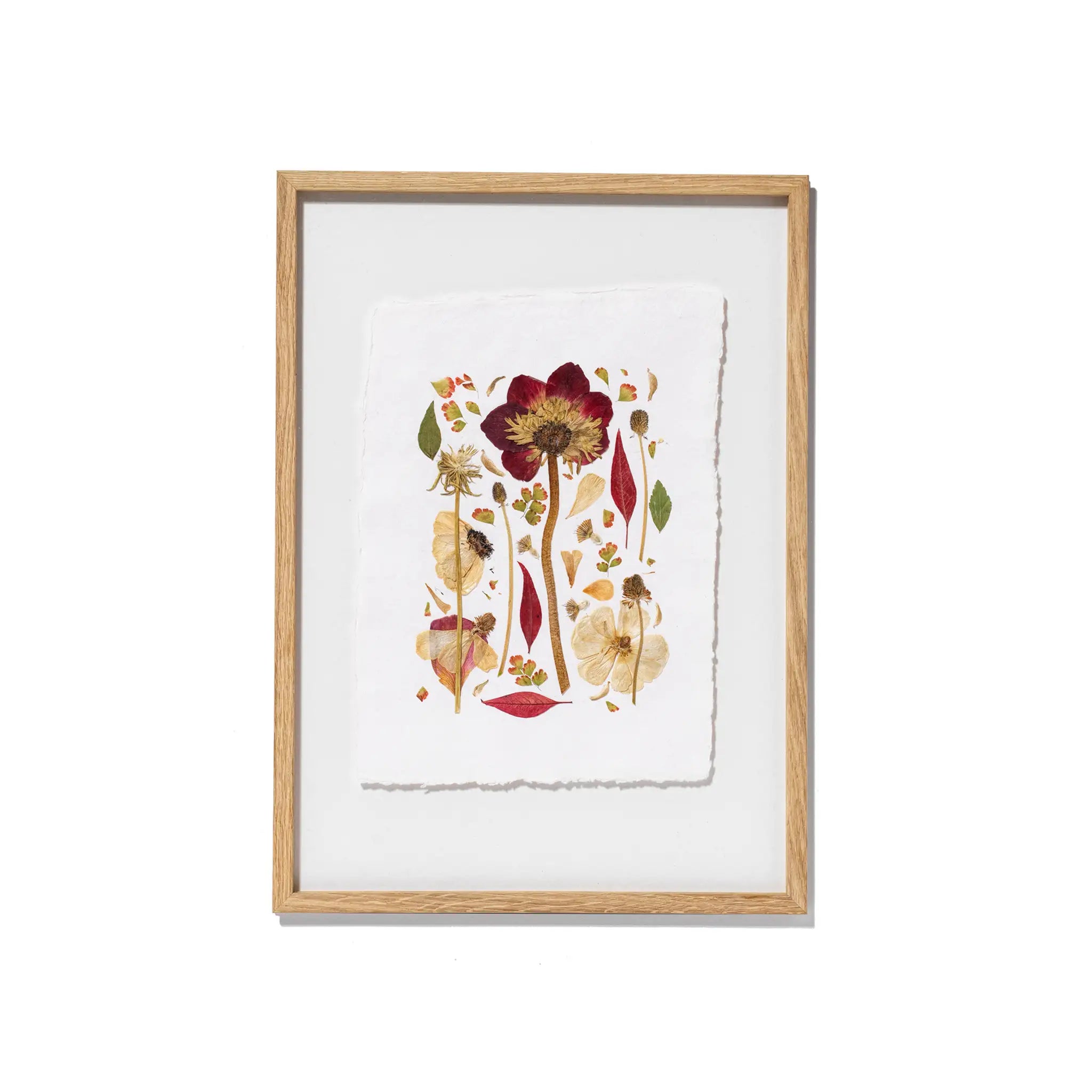 Framed botanical art featuring pressed flowers and leaves on textured white paper, set in a light natural wood frame against a clean white background.