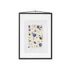 A framed floral art print featuring pressed pansies in purple, blue, and yellow on a white textured paper, enclosed in a black frame with a white background.