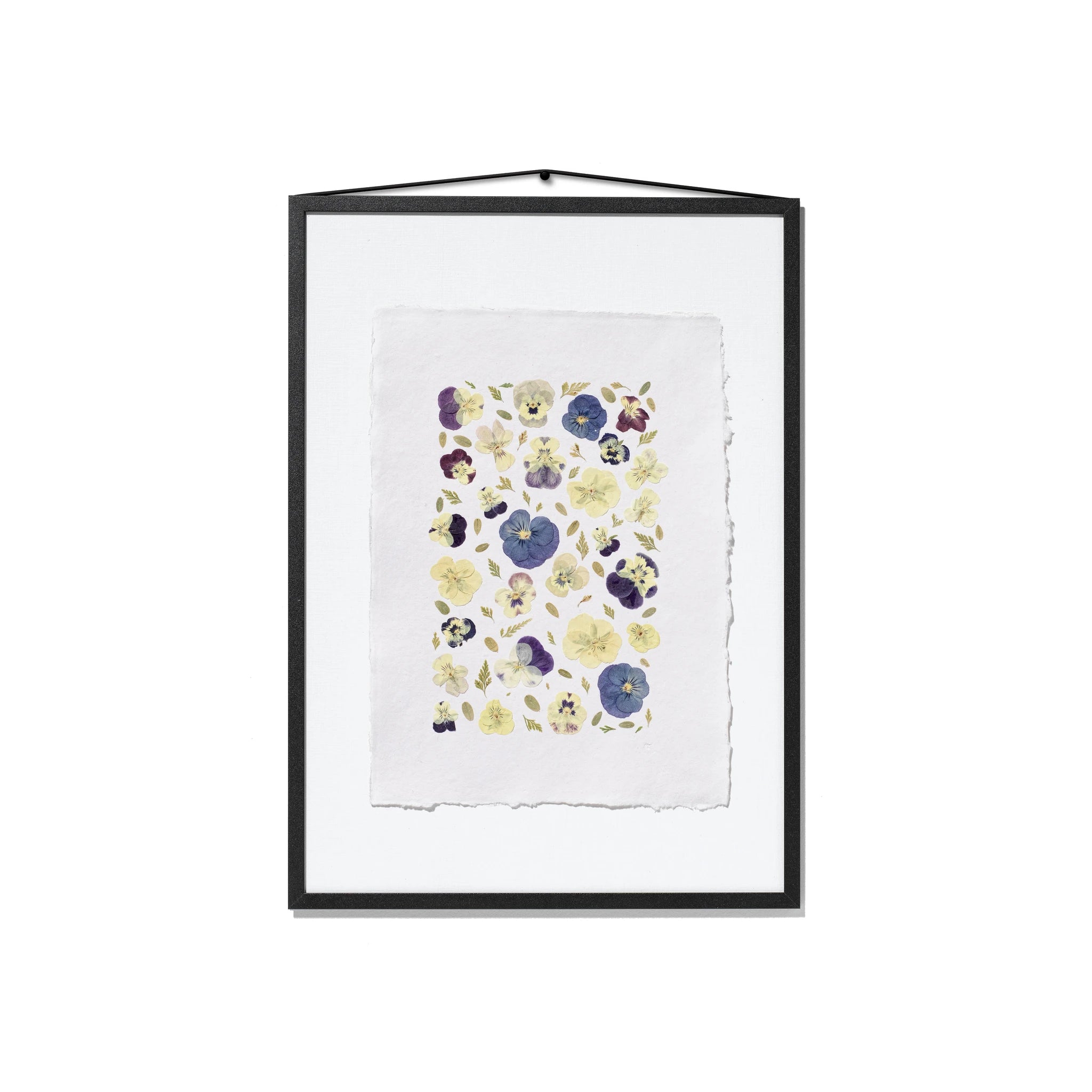 A framed floral art print featuring pressed pansies in purple, blue, and yellow on a white textured paper, enclosed in a black frame with a white background.