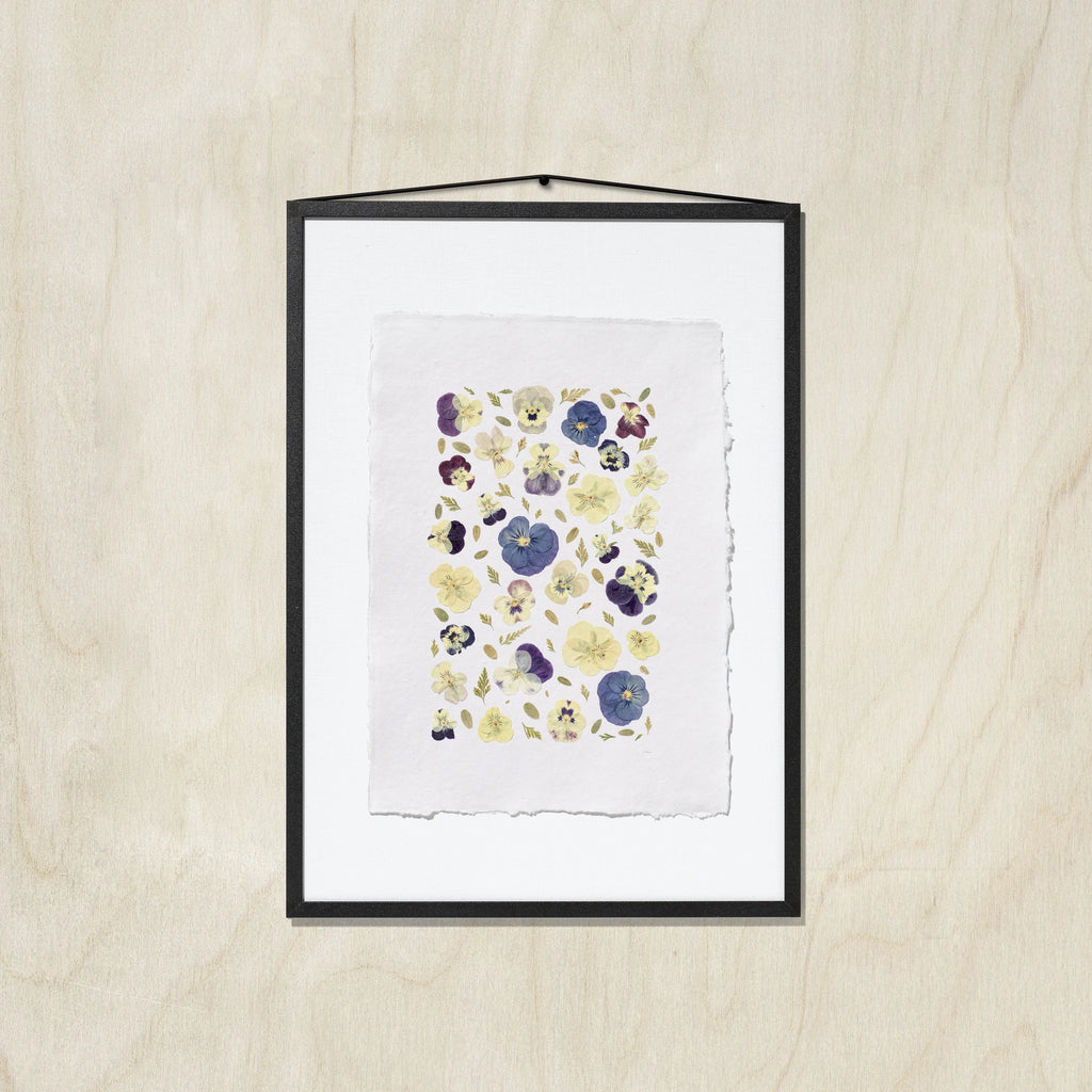 A framed floral art print featuring pressed or painted pansies in purple, blue, and yellow on a white textured paper background, enclosed in a black frame with a beige wooden background