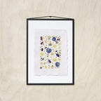 A framed floral art print featuring pressed or painted pansies in purple, blue, and yellow on a white textured paper background, enclosed in a black frame with a beige wooden background