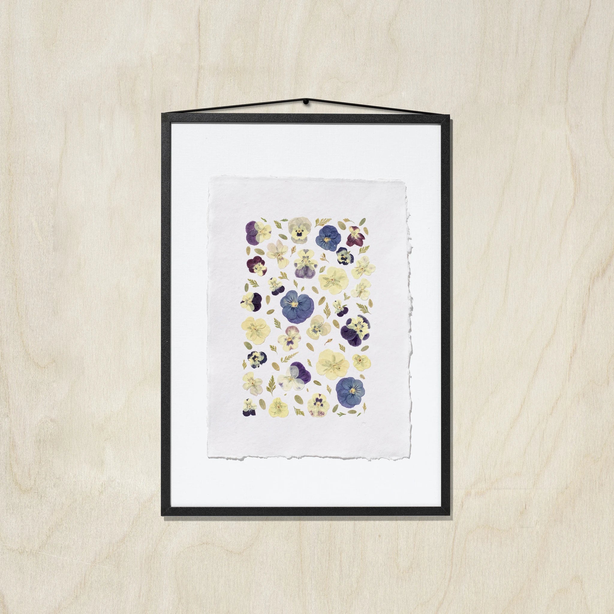 A framed floral art print featuring pressed or painted pansies in purple, blue, and yellow on a white textured paper background, enclosed in a black frame with a beige wooden background