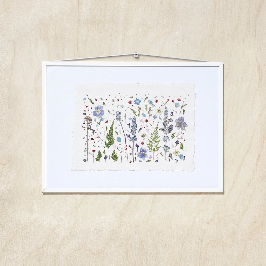 A framed botanical art featuring delicate flowers in shades of blue, purple, green, and red on textured white paper, presented in a white frame against a light wood background.