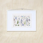 A framed botanical art featuring delicate flowers in shades of blue, purple, green, and red on textured white paper, presented in a white frame against a light wood background.