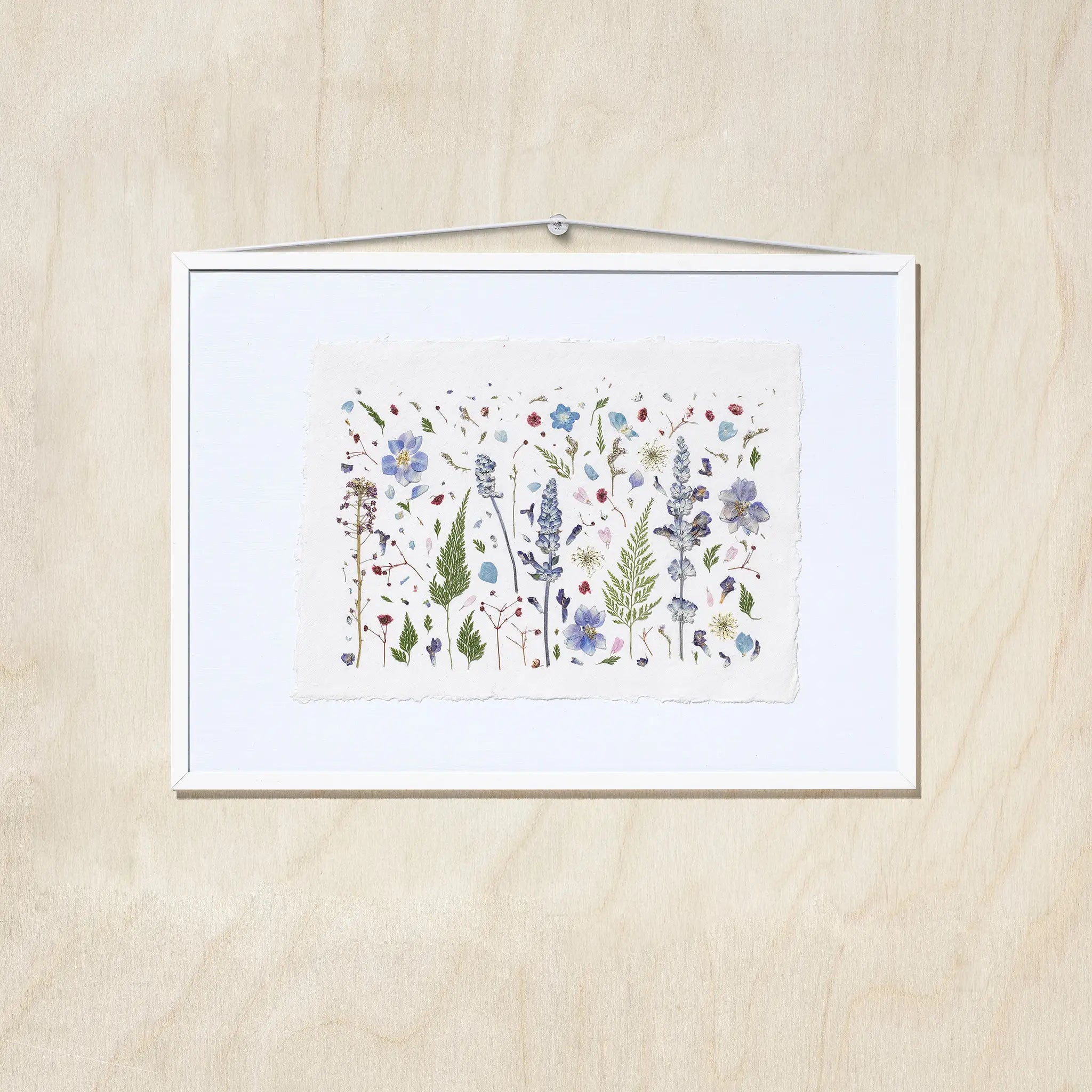A framed botanical art featuring delicate flowers in shades of blue, purple, green, and red on textured white paper, presented in a white frame against a light wood background.