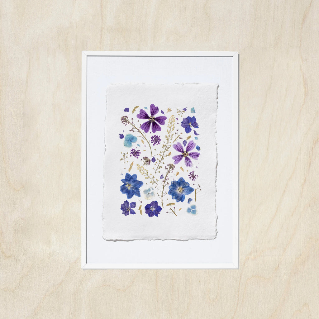 A framed floral art print featuring purple and blue pressed flowers on a textured white sheet of paper, set against a light oak wood background.