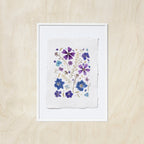 A framed floral art print featuring purple and blue pressed flowers on a textured white sheet of paper, set against a light oak wood background.
