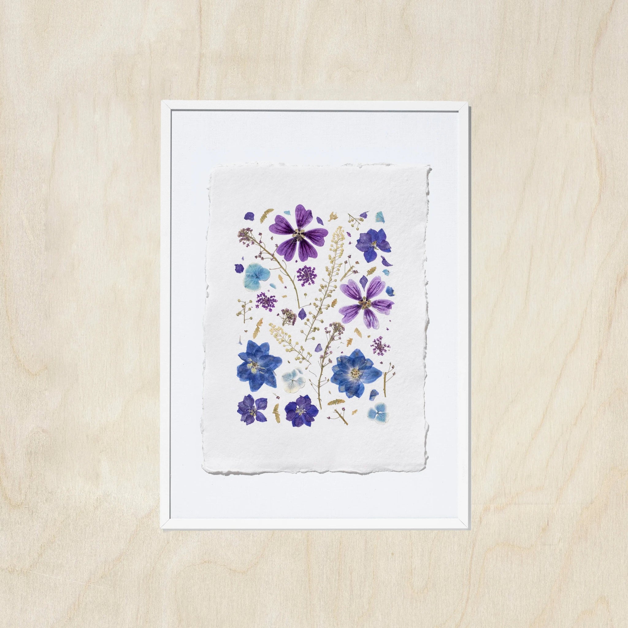 A framed floral art print featuring purple and blue pressed flowers on a textured white sheet of paper, set against a light oak wood background.