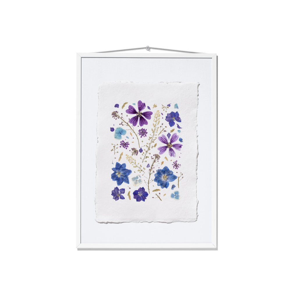 A framed botanical art print featuring pressed purple and blue flowers on a textured white paper background, housed in a white frame with a hanging wire at the top.