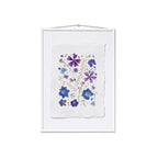 A framed botanical art print featuring pressed purple and blue flowers on a textured white paper background, housed in a white frame with a hanging wire at the top.