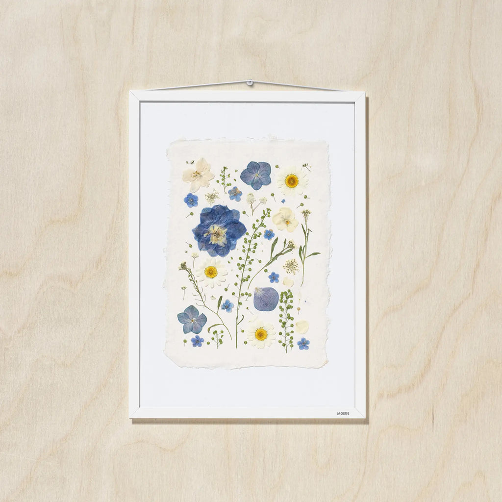 A framed botanical art featuring blue, yellow, and cream flowers on a textured off-white paper background, mounted on a light oak wood wall.