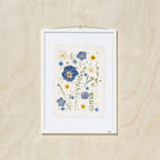 A framed botanical art featuring blue, yellow, and cream flowers on a textured off-white paper background, mounted on a light oak wood wall.