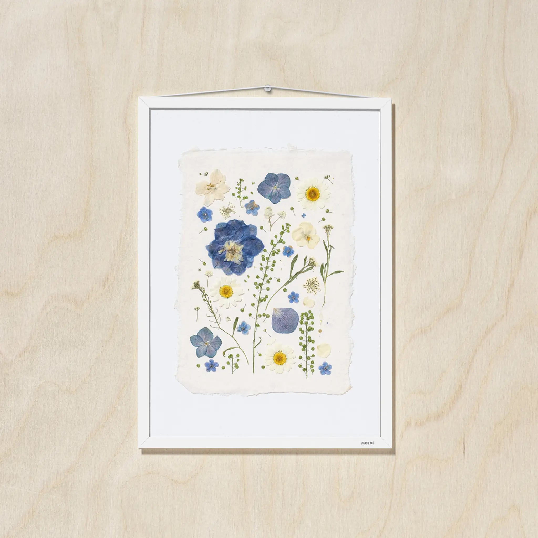 A framed botanical art featuring blue, yellow, and cream flowers on a textured off-white paper background, mounted on a light oak wood wall.
