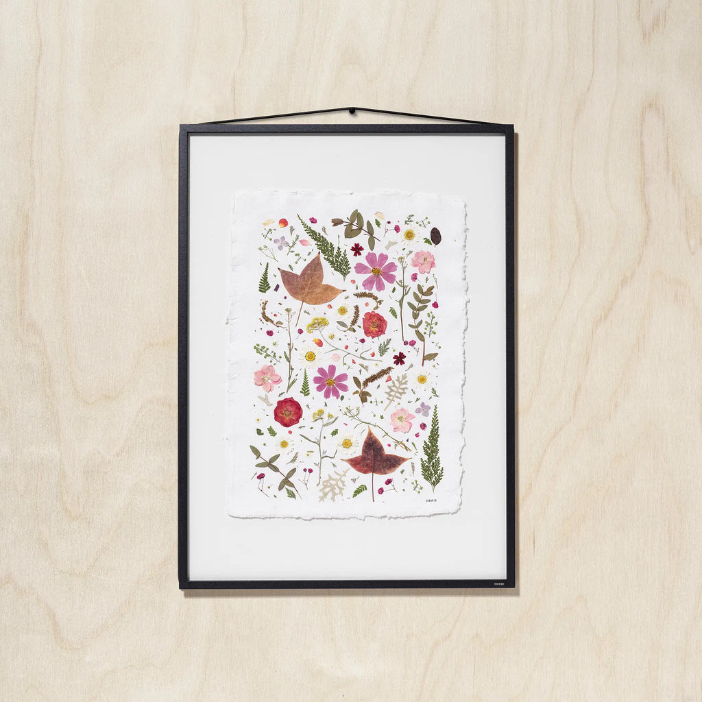 A framed botanical art featuring colorful pressed flowers, leaves on textured white paper, hung on a light wood wall.