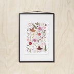 A framed botanical art featuring colorful pressed flowers, leaves on textured white paper, hung on a light wood wall.