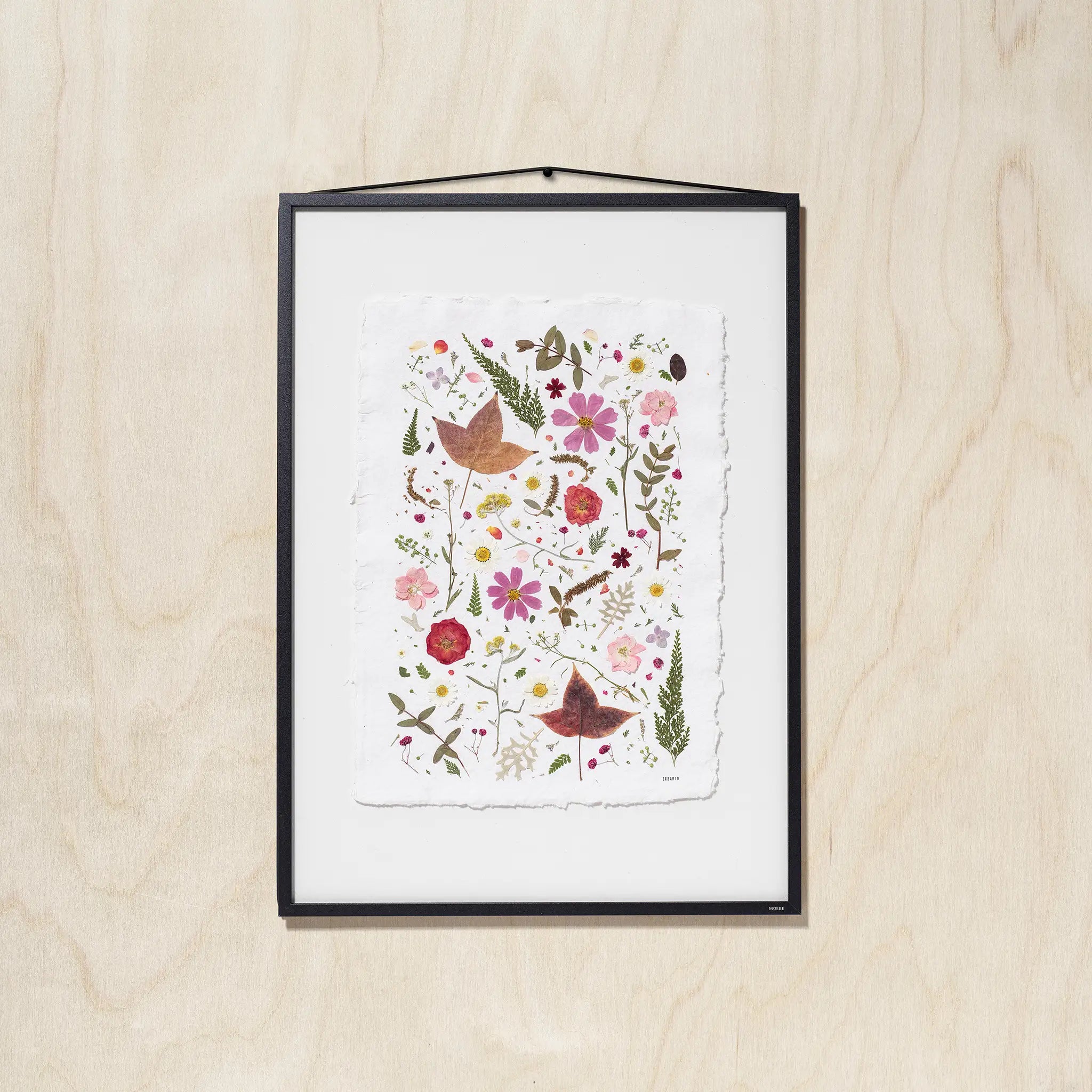 A framed botanical art featuring colorful pressed flowers, leaves on textured white paper, hung on a light wood wall.