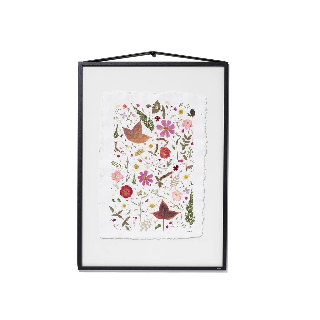 A framed botanical art featuring pressed flowers and leaves on a white textured paper background, housed in a black frame with a hanging cord.