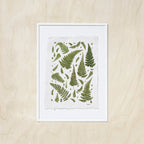 A framed botanical art print featuring green fern leaves on a textured off-white sheet, in a white frame, displayed against a light oak wood background.