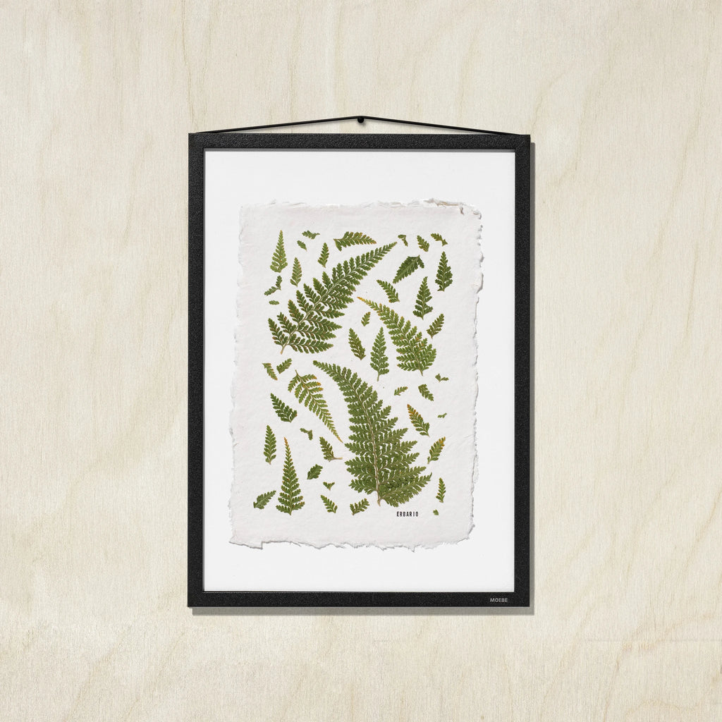 A framed botanical art print featuring green fern leaves on a textured off-white background, presented in a black frame against a light wood wall.