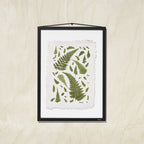 A framed botanical art print featuring green fern leaves on a textured off-white background, presented in a black frame against a light wood wall.