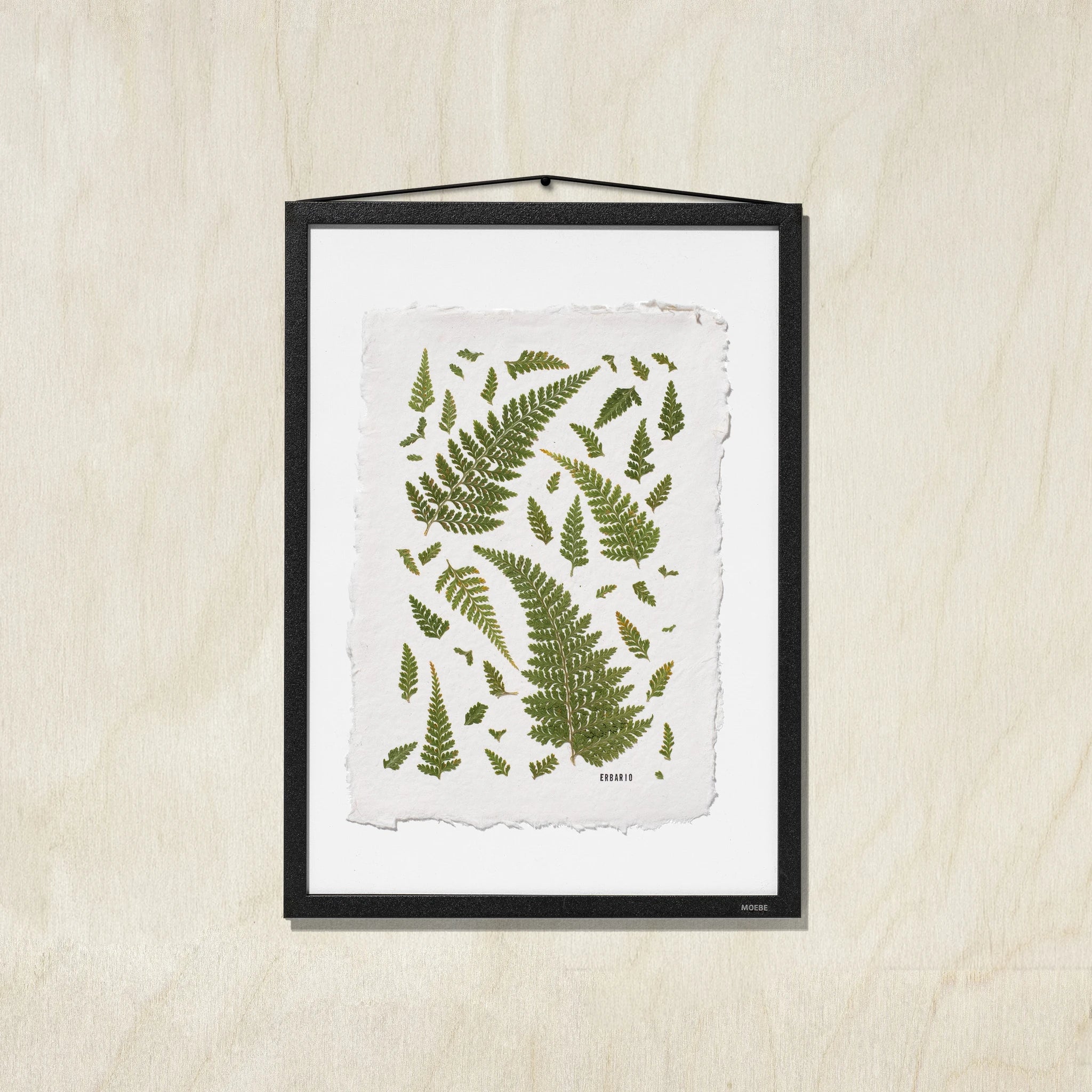 A framed botanical art print featuring green fern leaves on a textured off-white background, presented in a black frame against a light wood wall.
