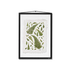 A framed botanical art print featuring green fern leaves on a textured off-white sheet, presented in a black frame with a hanging cord against a white background.