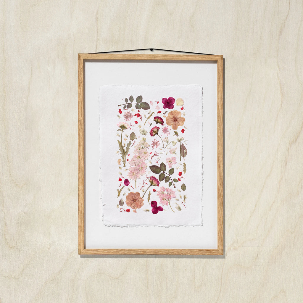 A framed botanical art print featuring delicate flowers and leaves in soft pink, red, beige, and green tones, set in an oak wood frame with a white background, hanging against an oak wall.
