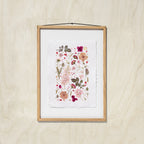 A framed botanical art print featuring delicate flowers and leaves in soft pink, red, beige, and green tones, set in an oak wood frame with a white background, hanging against an oak wall.