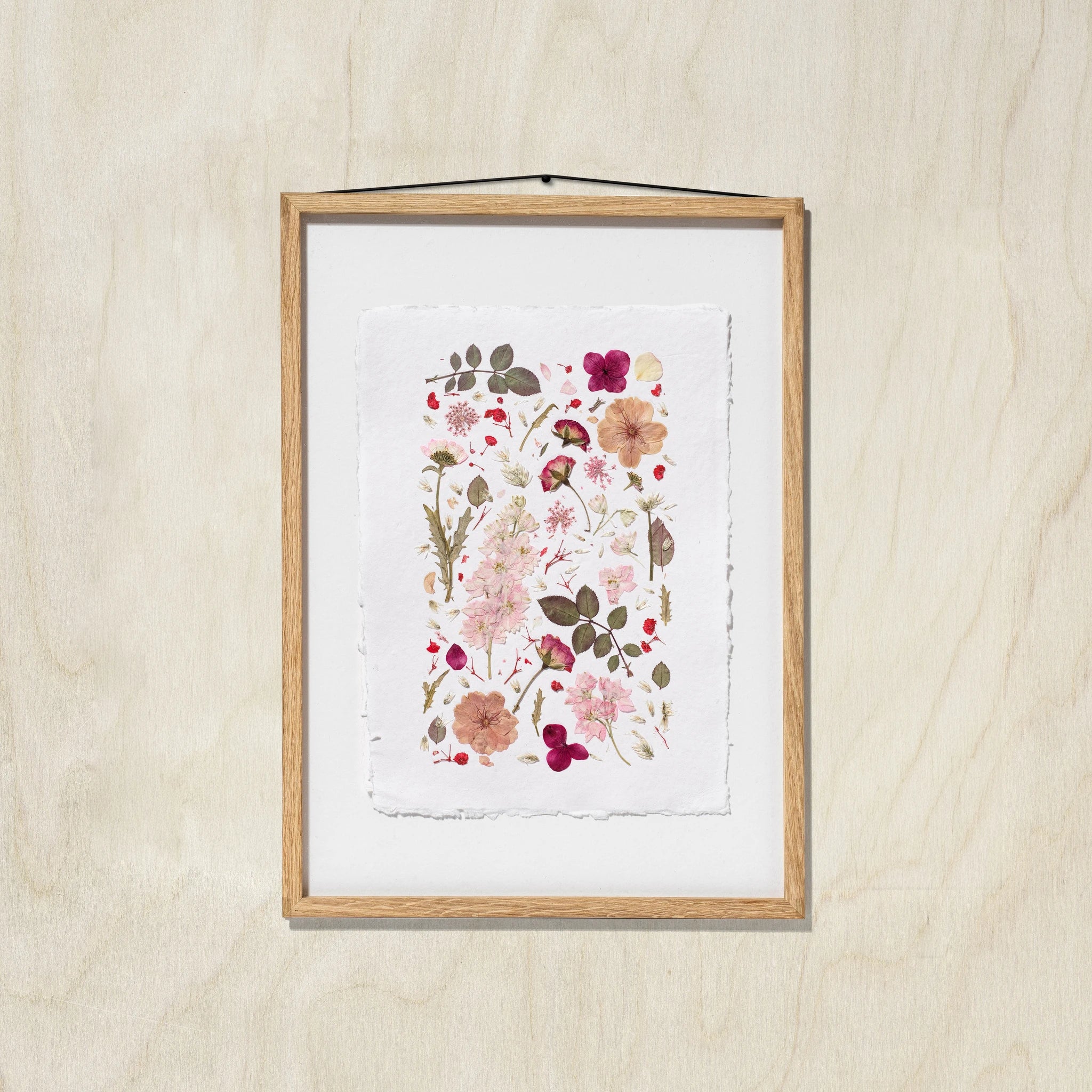 A framed botanical art print featuring delicate flowers and leaves in soft pink, red, beige, and green tones, set in an oak wood frame with a white background, hanging against an oak wall.